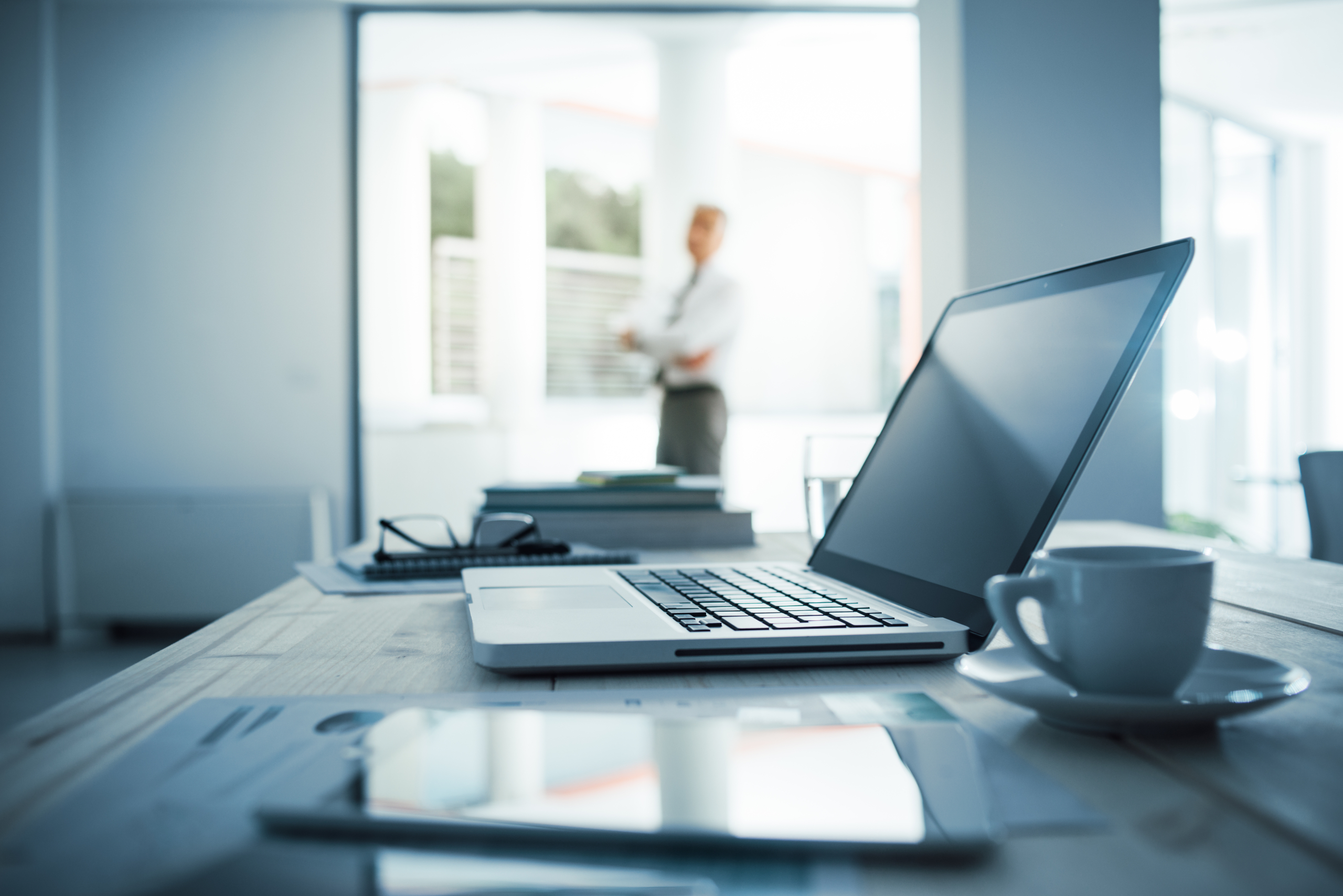 Businessman standing with arms crossed in his office, desktop with laptop on foreground, selective focus
